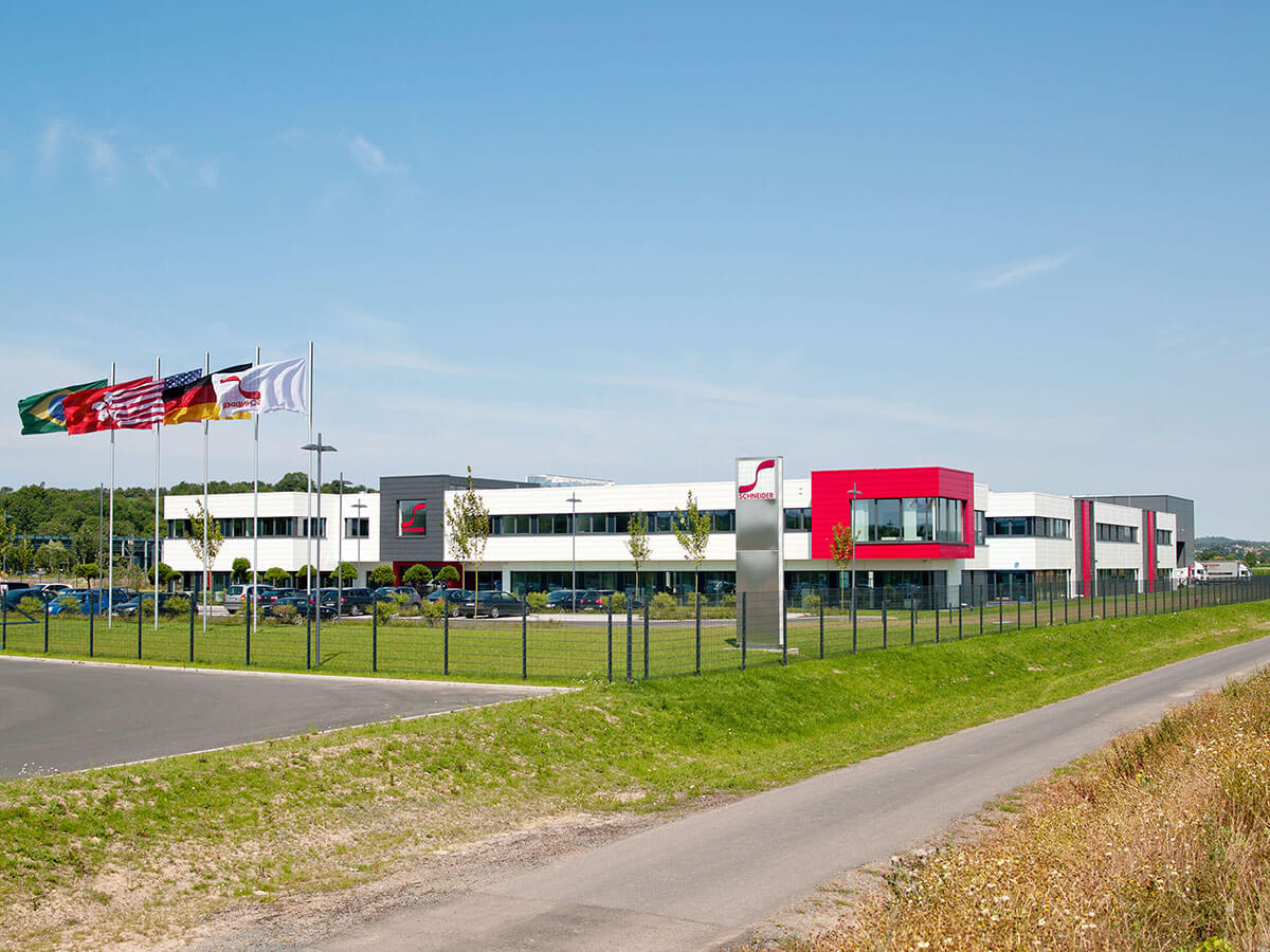 Fahnen wehen vor modernem Bürogebäude auf grüner Wiese unter blauem Himmel.