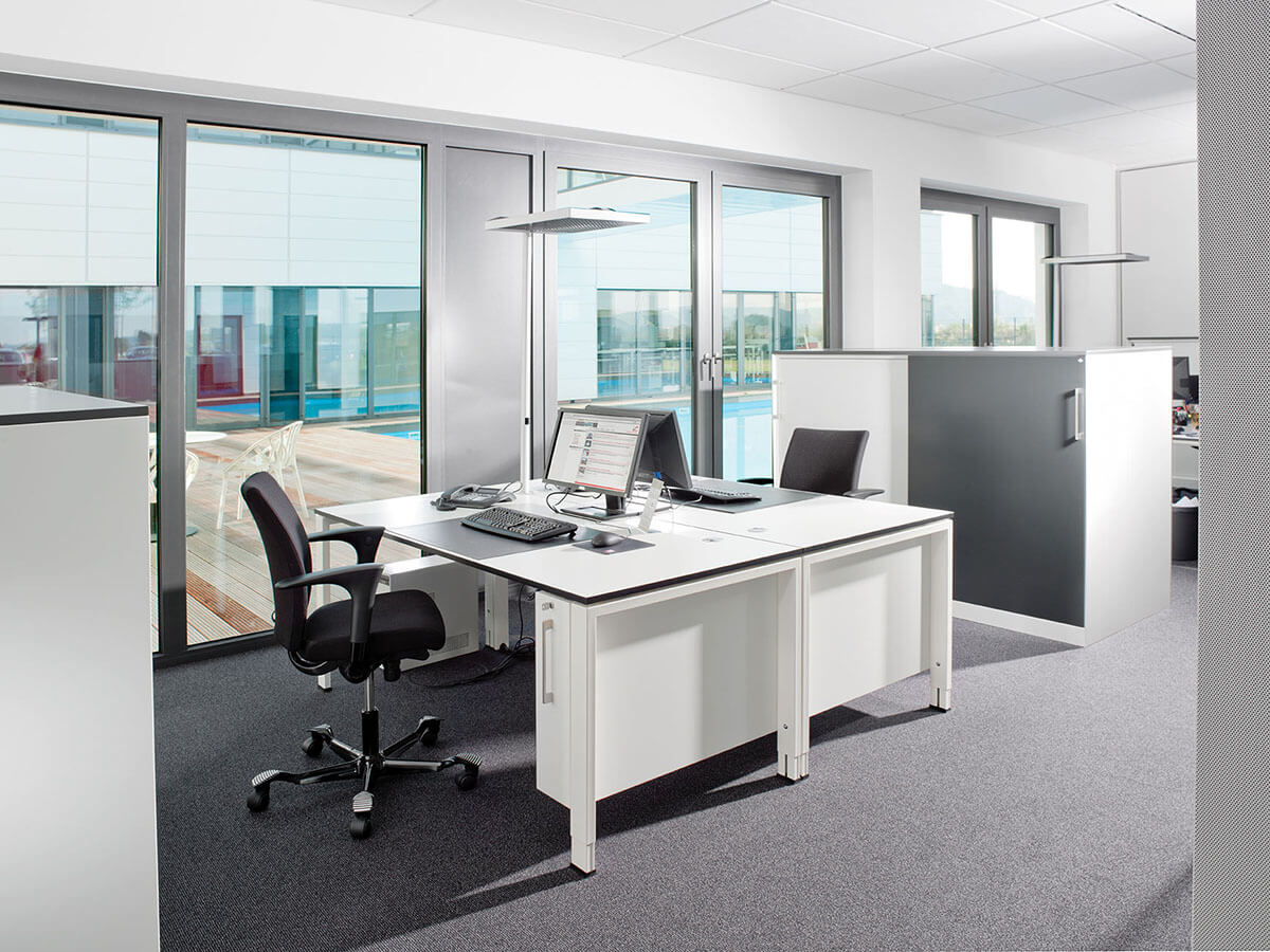 Bürotisch mit Computerarbeitsplatz in modernem Büro vor großen Fenstern.