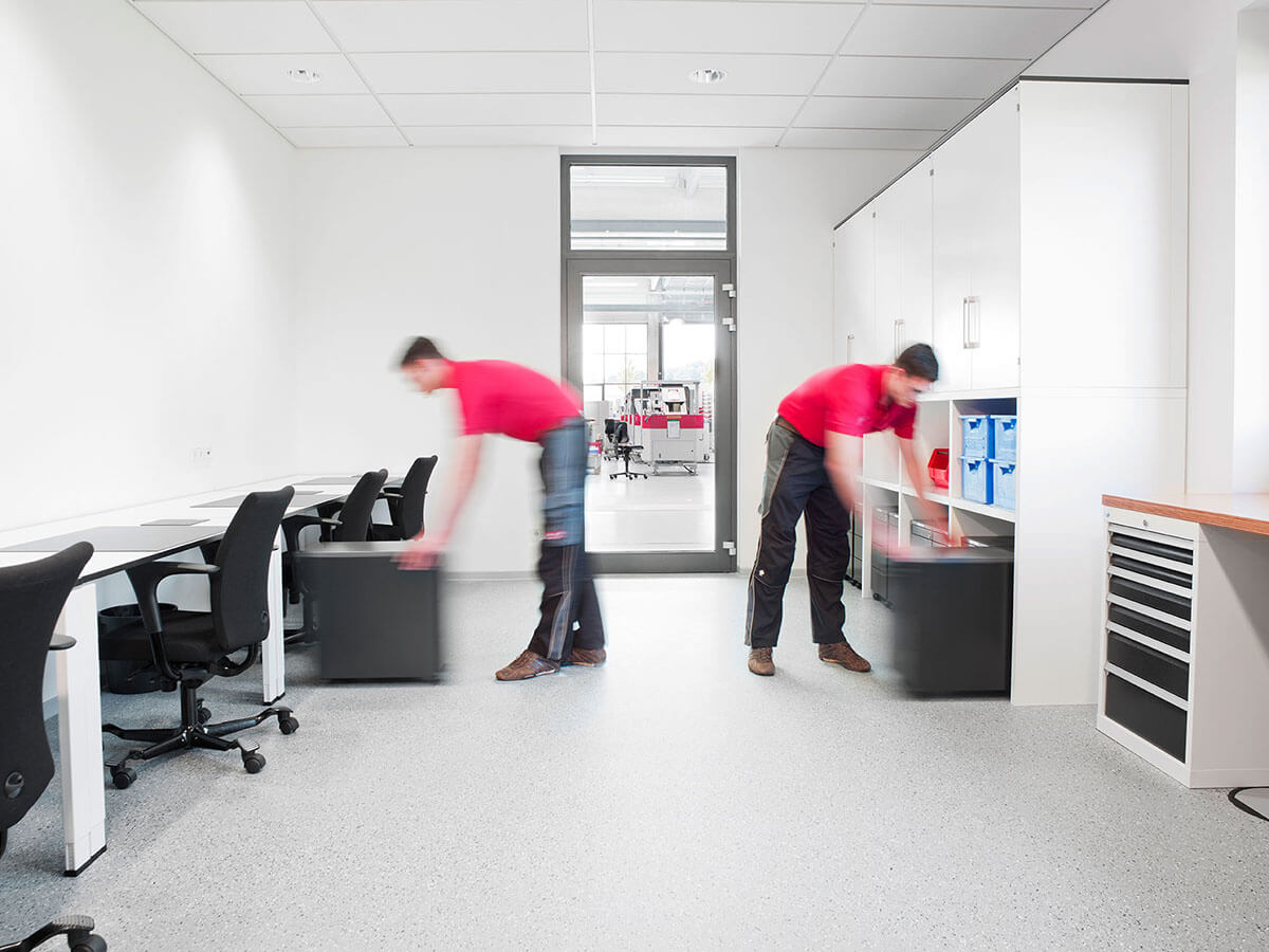 Zwei Männer bewegen Aktenschränke in einem Büro mit Schreibtischen und Stühlen.