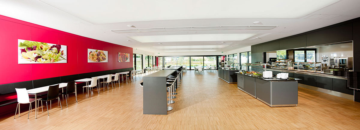 Leere Kantine mit Esstischen und Buffet, helle Holzböden, rote Wanddeko.
